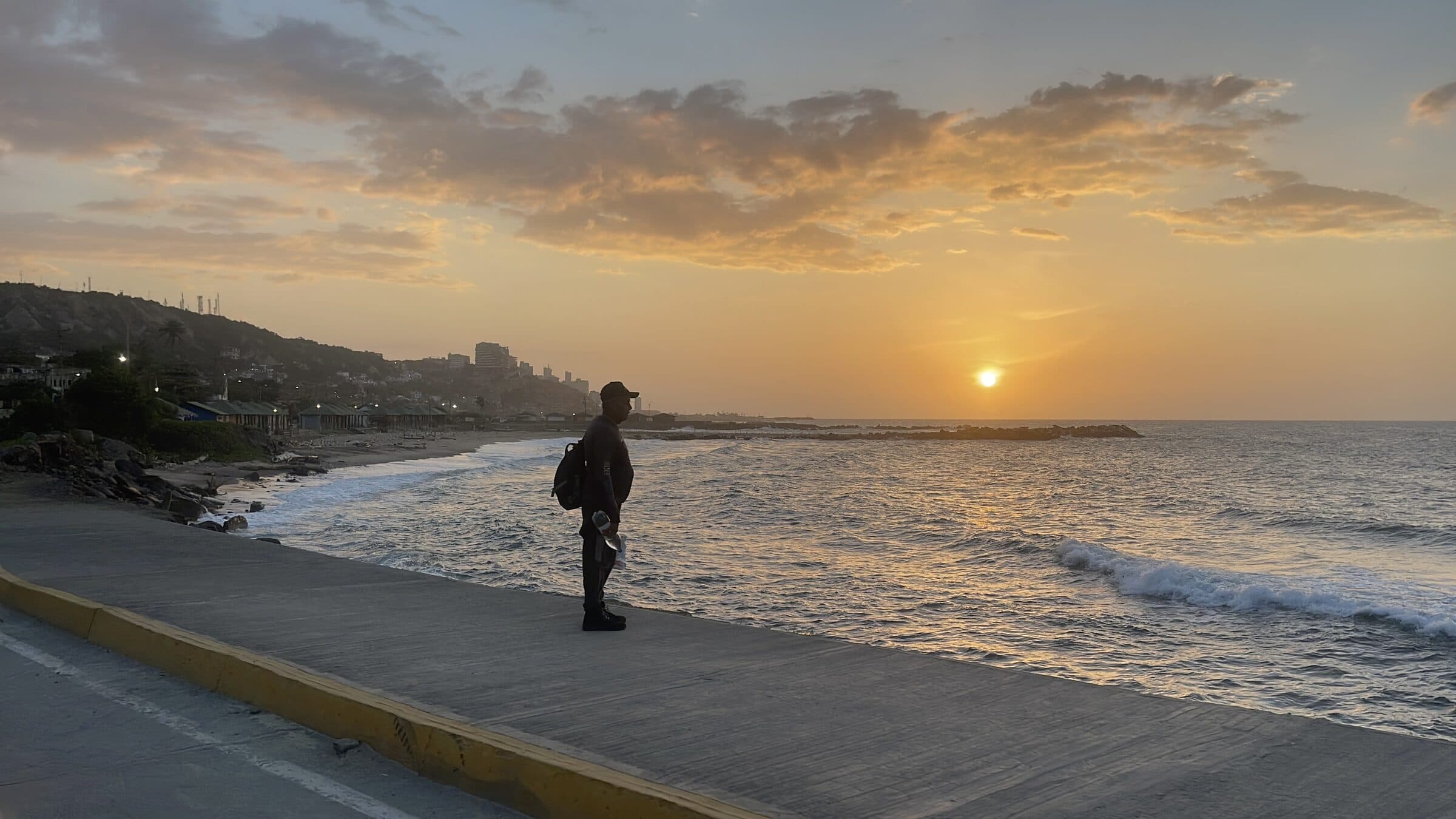 Sunset over the La Guaira coastline in Venezuela, with a silhouetted figure standing on the seafront promenade.