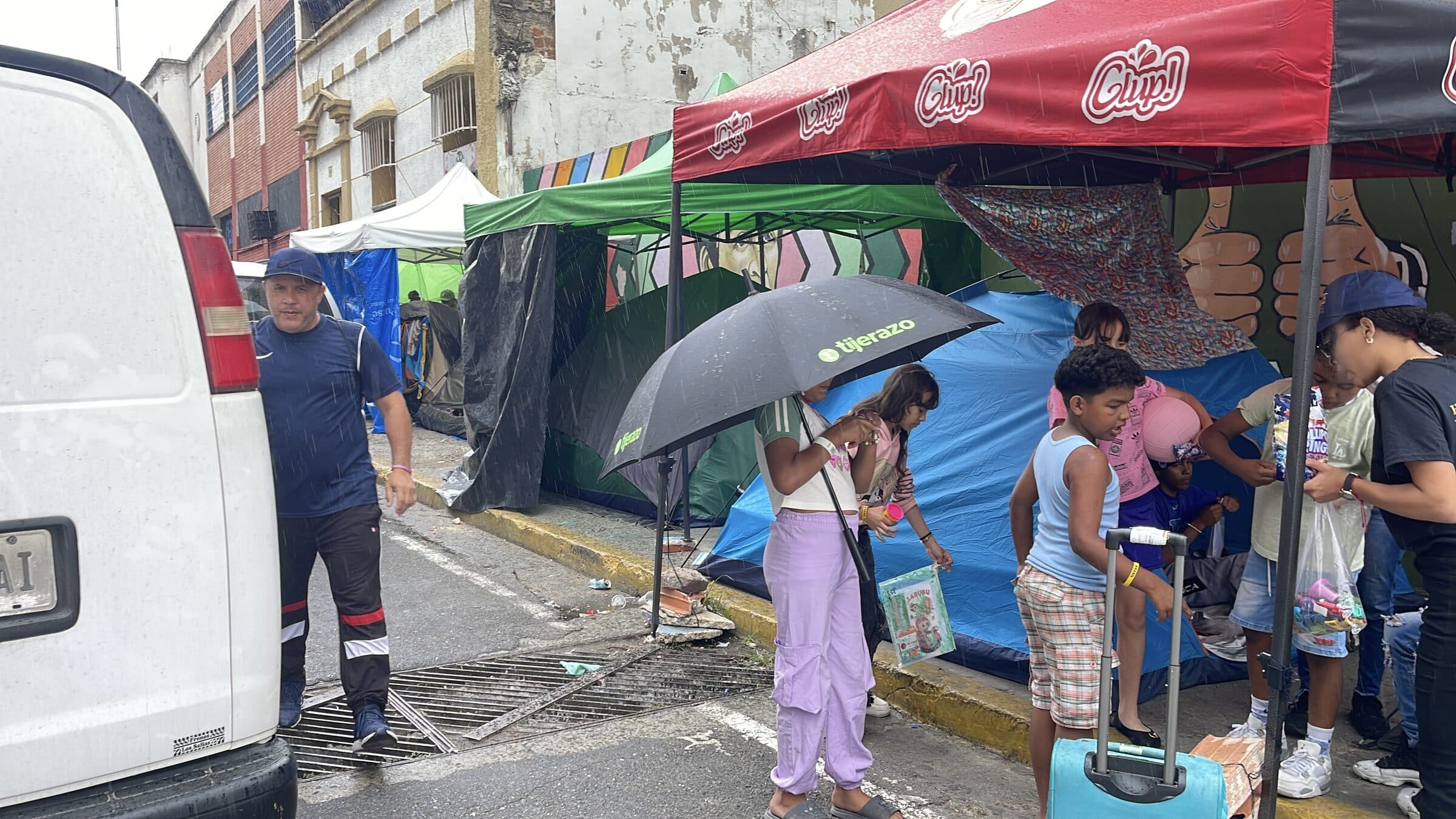 Wide view of the tent camp on a rainy day, with an IHE volunteer distributing supplies under a canopy.