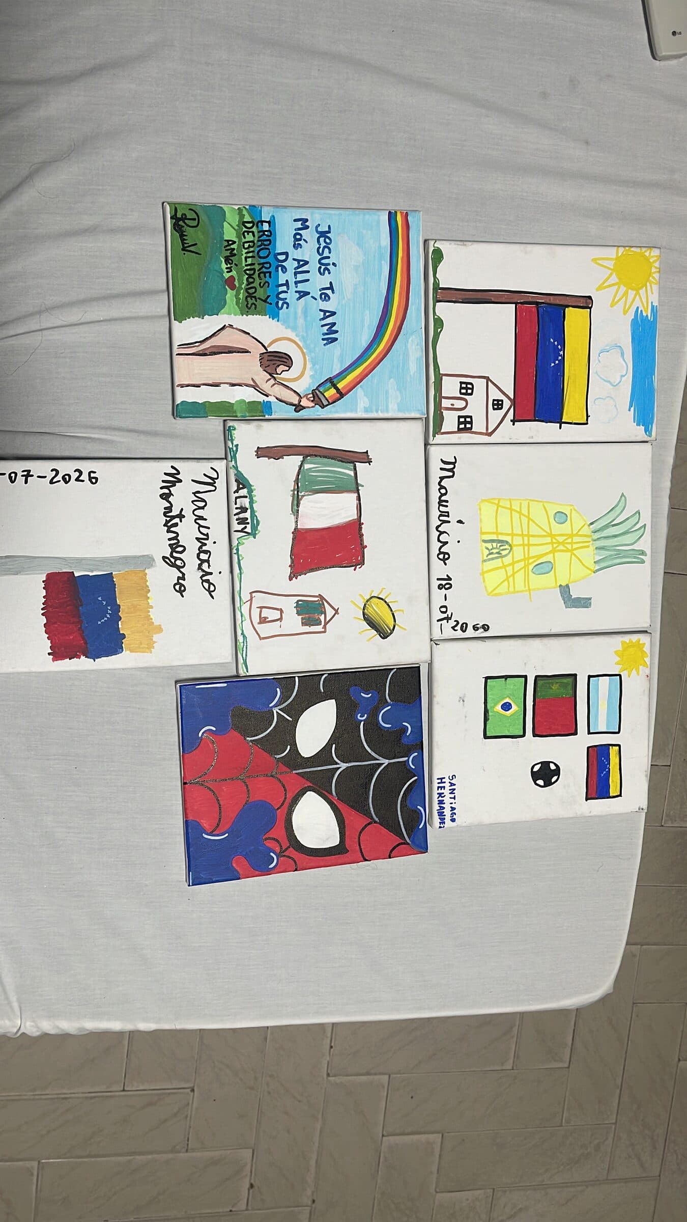 Seven finished canvases from the children's art workshop laid out together, showing flags, a pineapple and other drawings dated July 18, 2026.