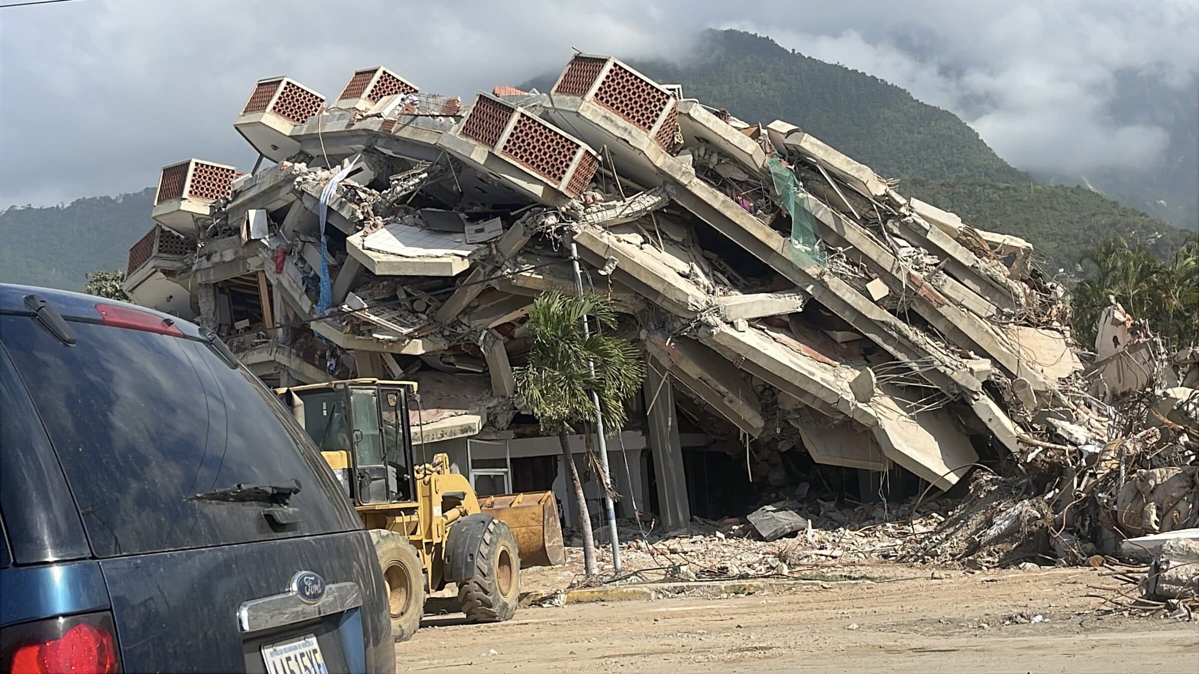 Multi-story building pancaked by the earthquake in La Guaira, with a front loader clearing debris at its base.
