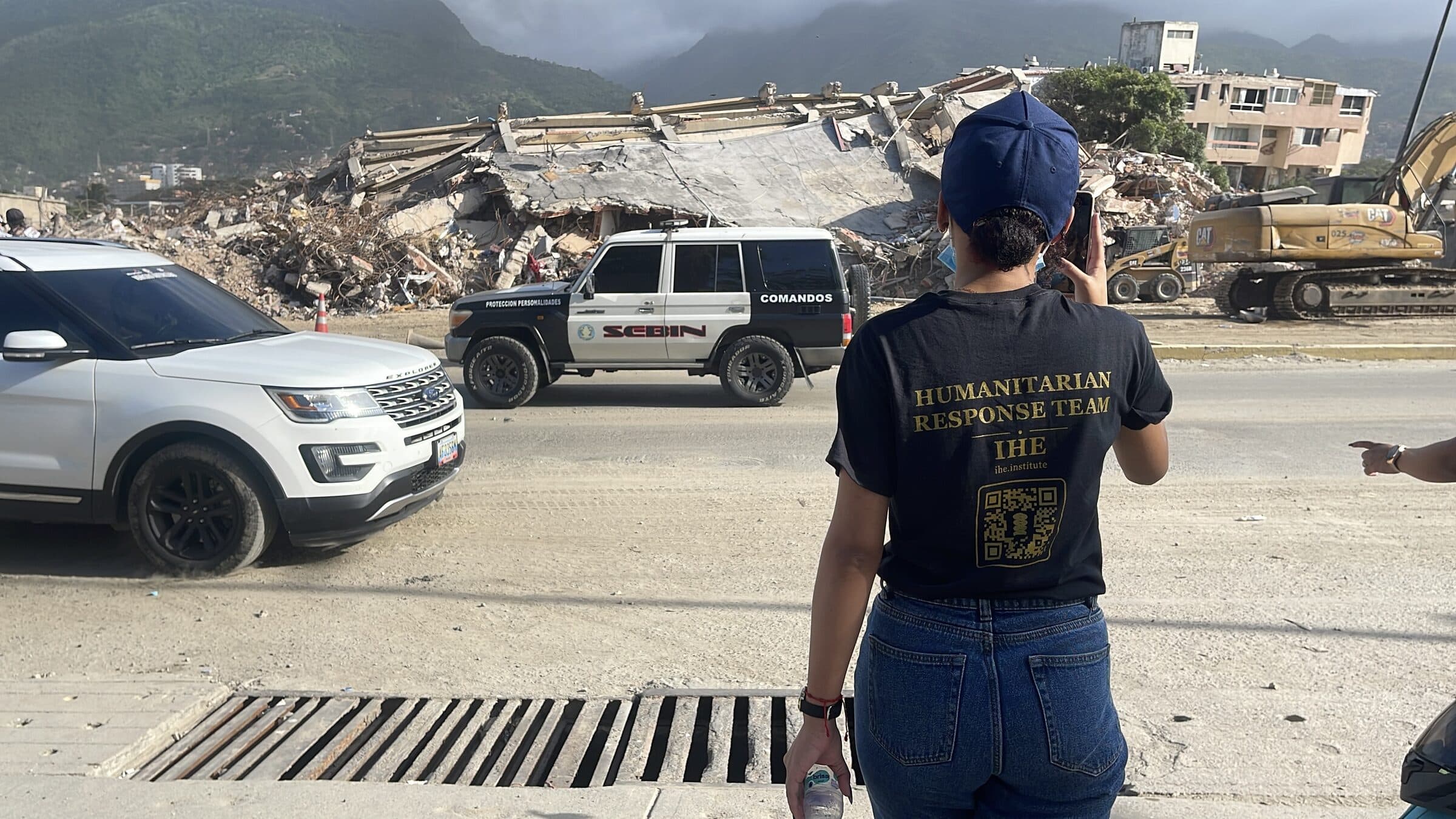 IHE Humanitarian Response Team member documenting the earthquake-collapsed buildings in La Guaira, Venezuela, with heavy machinery working in the background.
