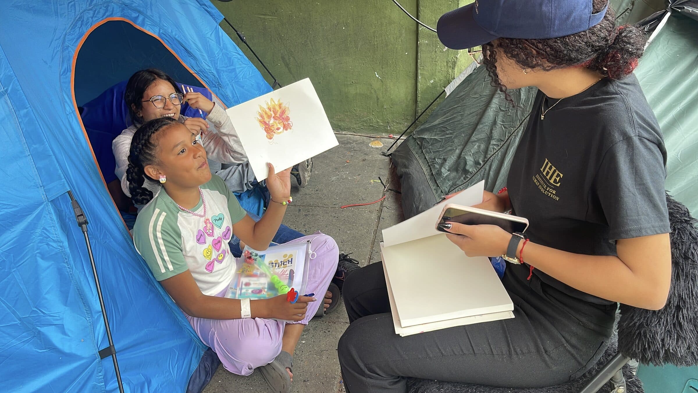 A girl smiles widely as she shows her drawing to the camera during an IHE art workshop.