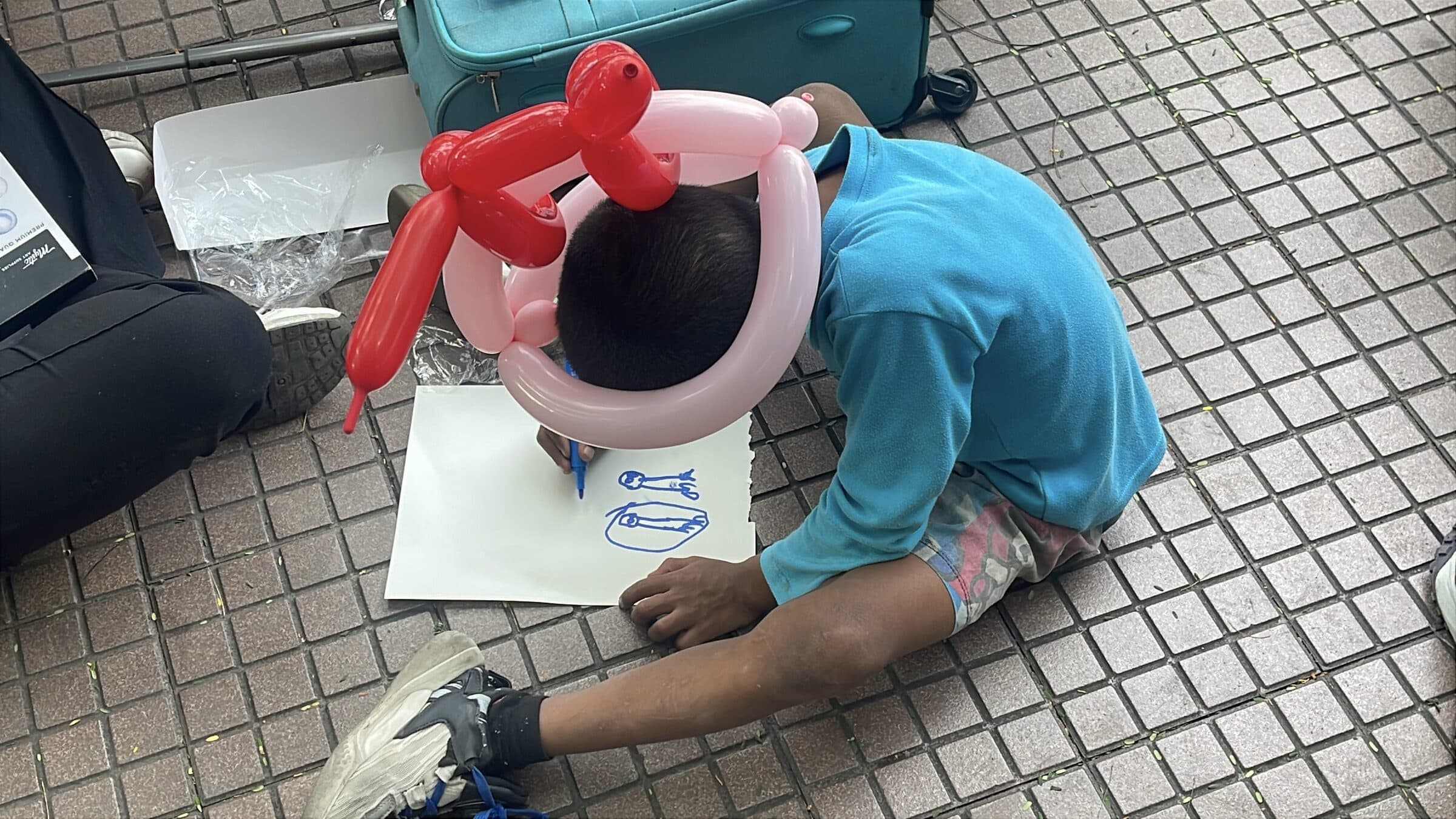 A boy wearing a balloon hat leans over his drawing on the ground, fully absorbed in coloring; his face is not visible.