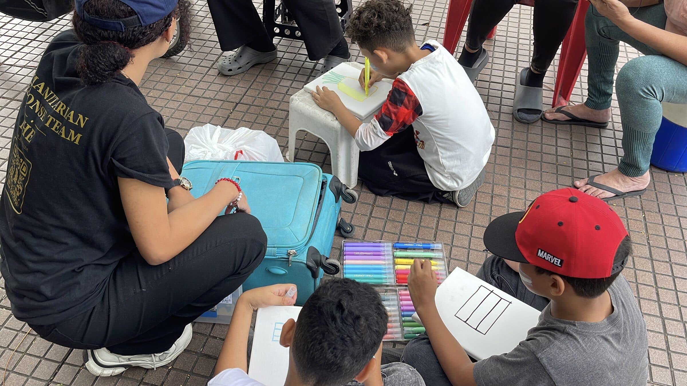 Seen from behind, a boy paints a pineapple on a small canvas during an IHE art workshop; a volunteer's Humanitarian Response Team shirt is in the foreground.