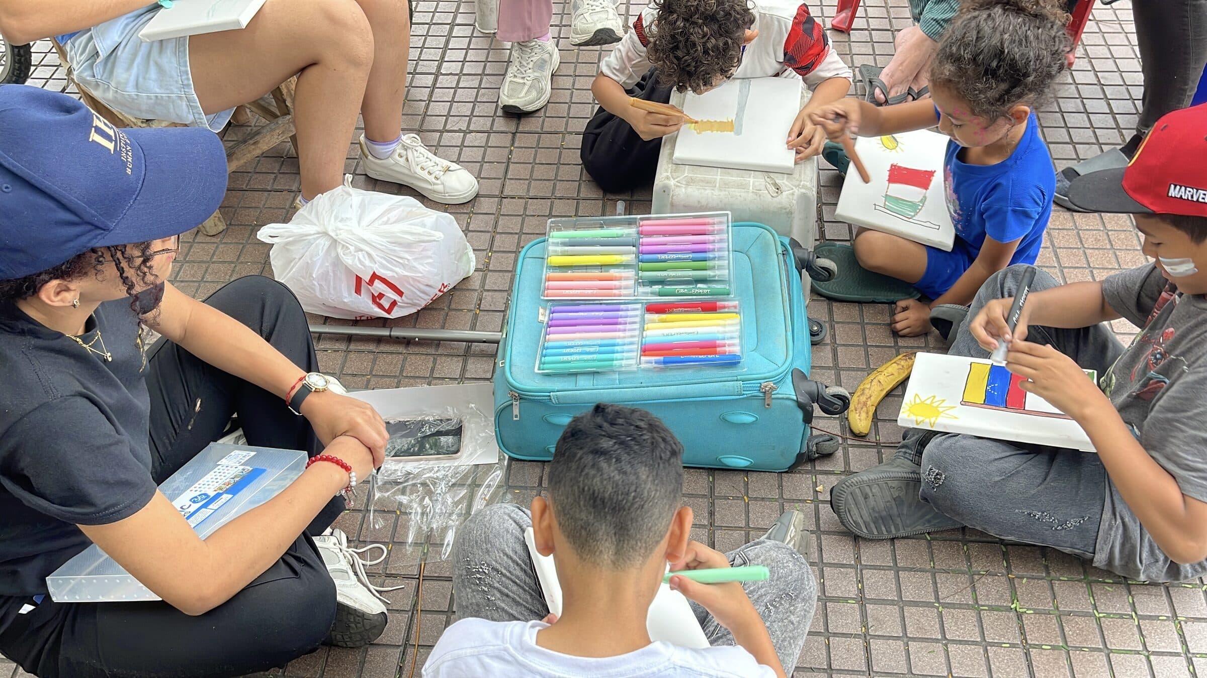 Children paint flags, including Venezuela's, on small canvases around a suitcase used as a table full of marker sets.