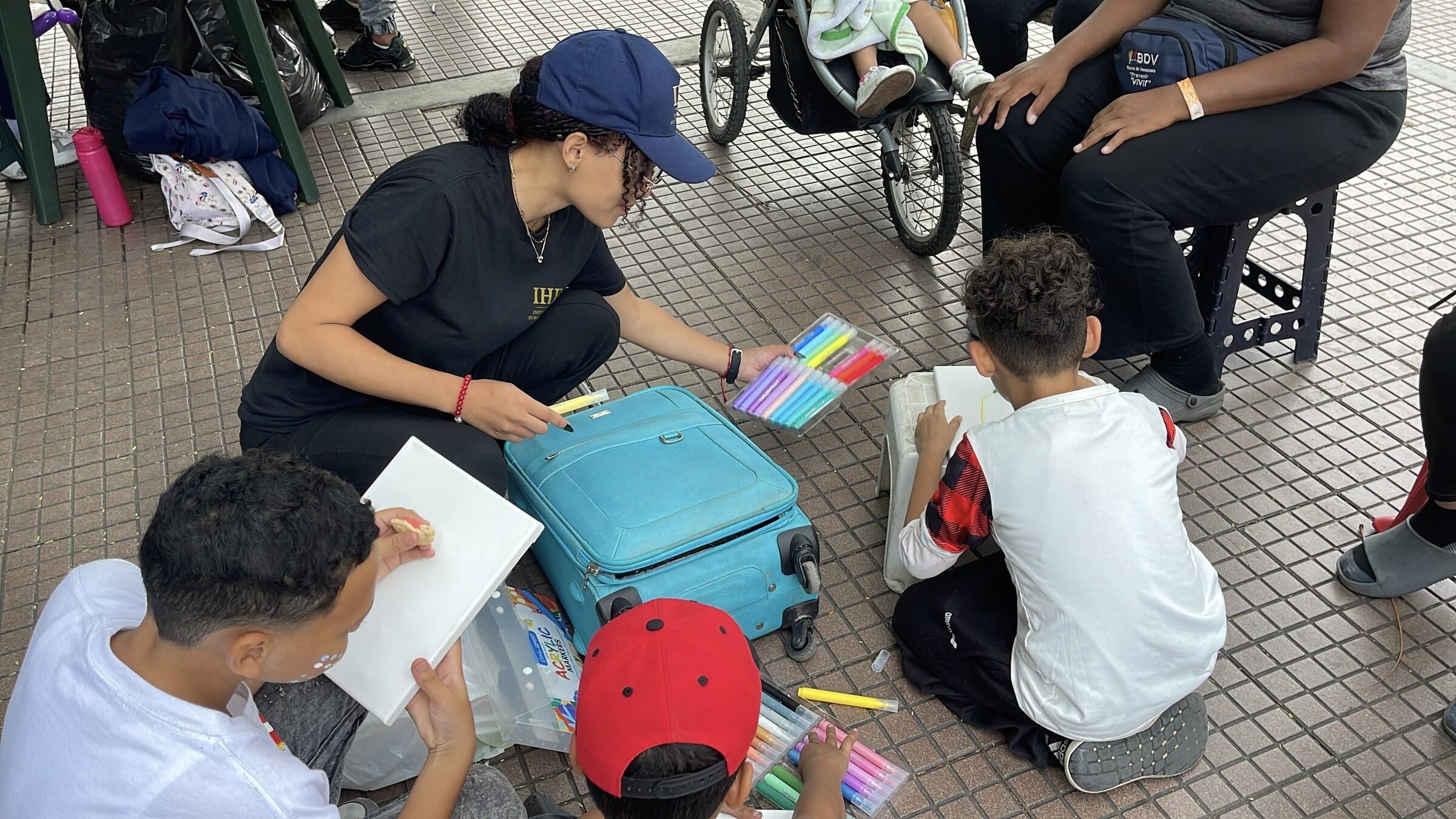 An IHE volunteer offers a tray of colored markers to children painting canvases at an outdoor art workshop in a displaced families camp in Venezuela.