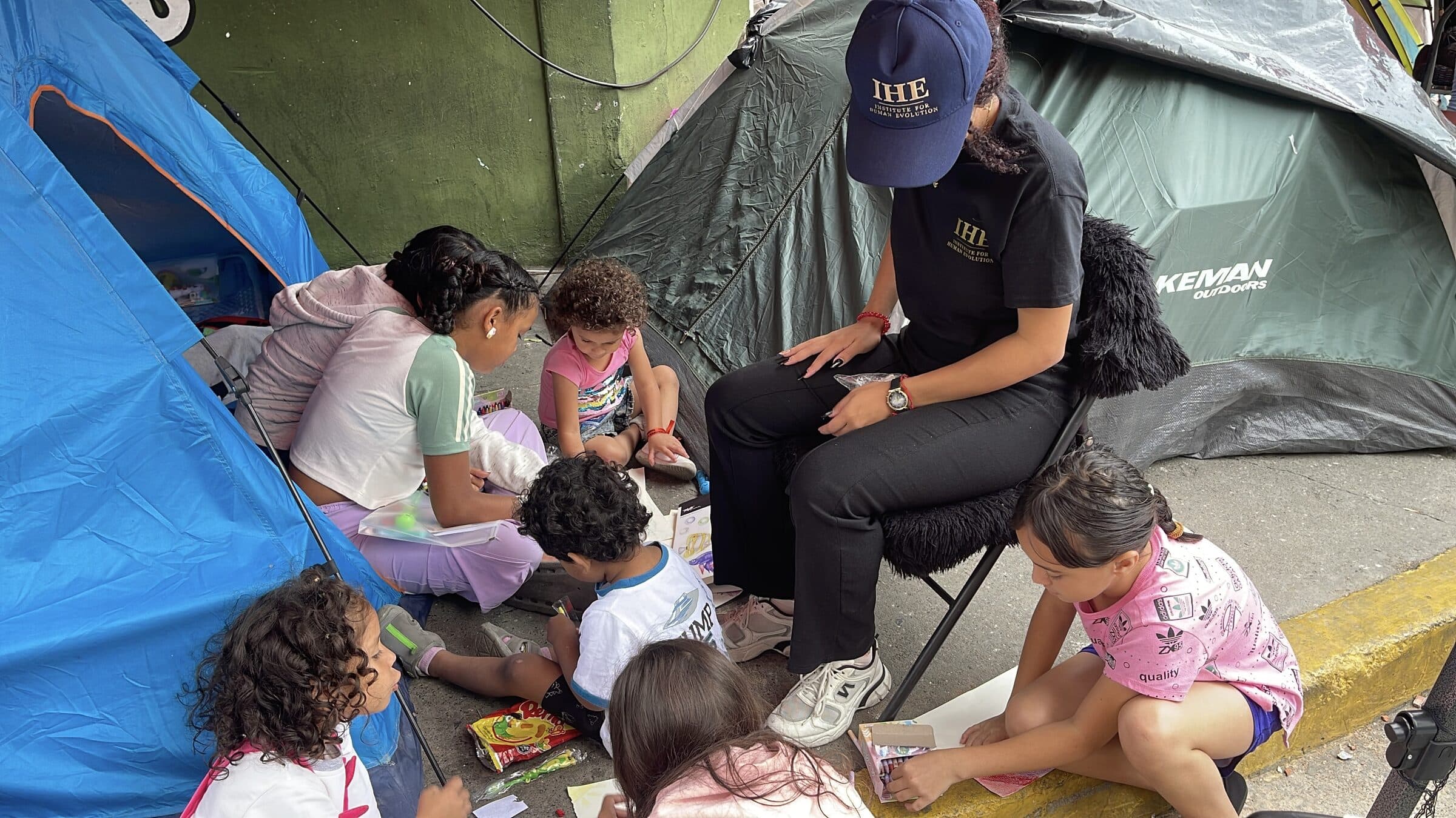 An IHE volunteer sits among children absorbed in drawing between tents at the camp.