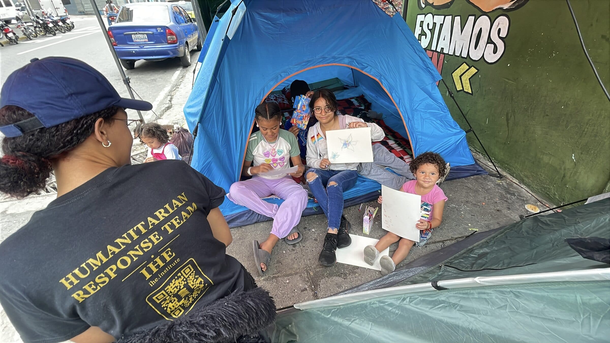 Seen from above, children's hands draw on paper at the curb while an IHE volunteer sits with them.