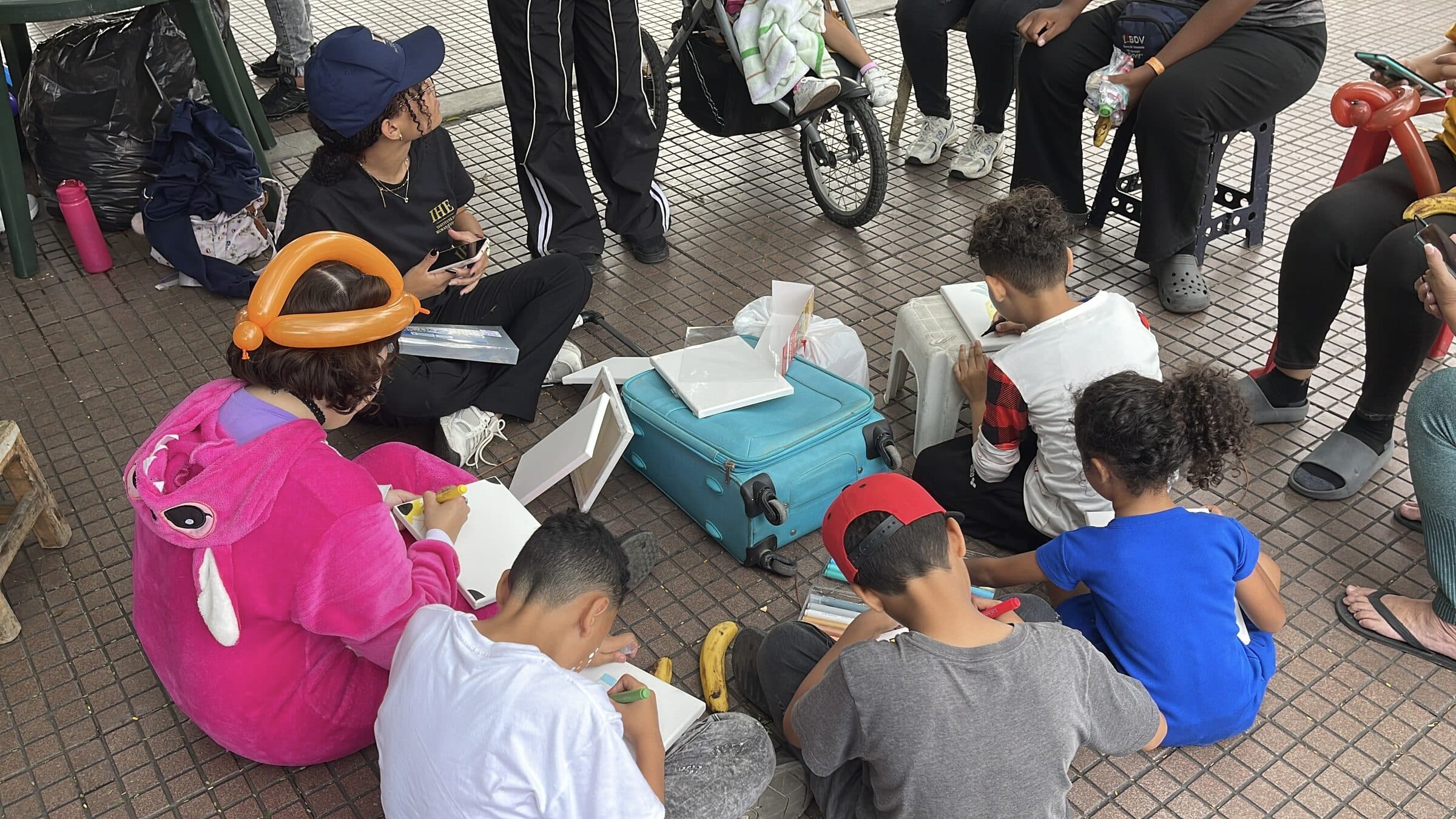 Wide view of an outdoor art workshop: children seen from behind paint canvases around a suitcase of art supplies while families gather in a circle.