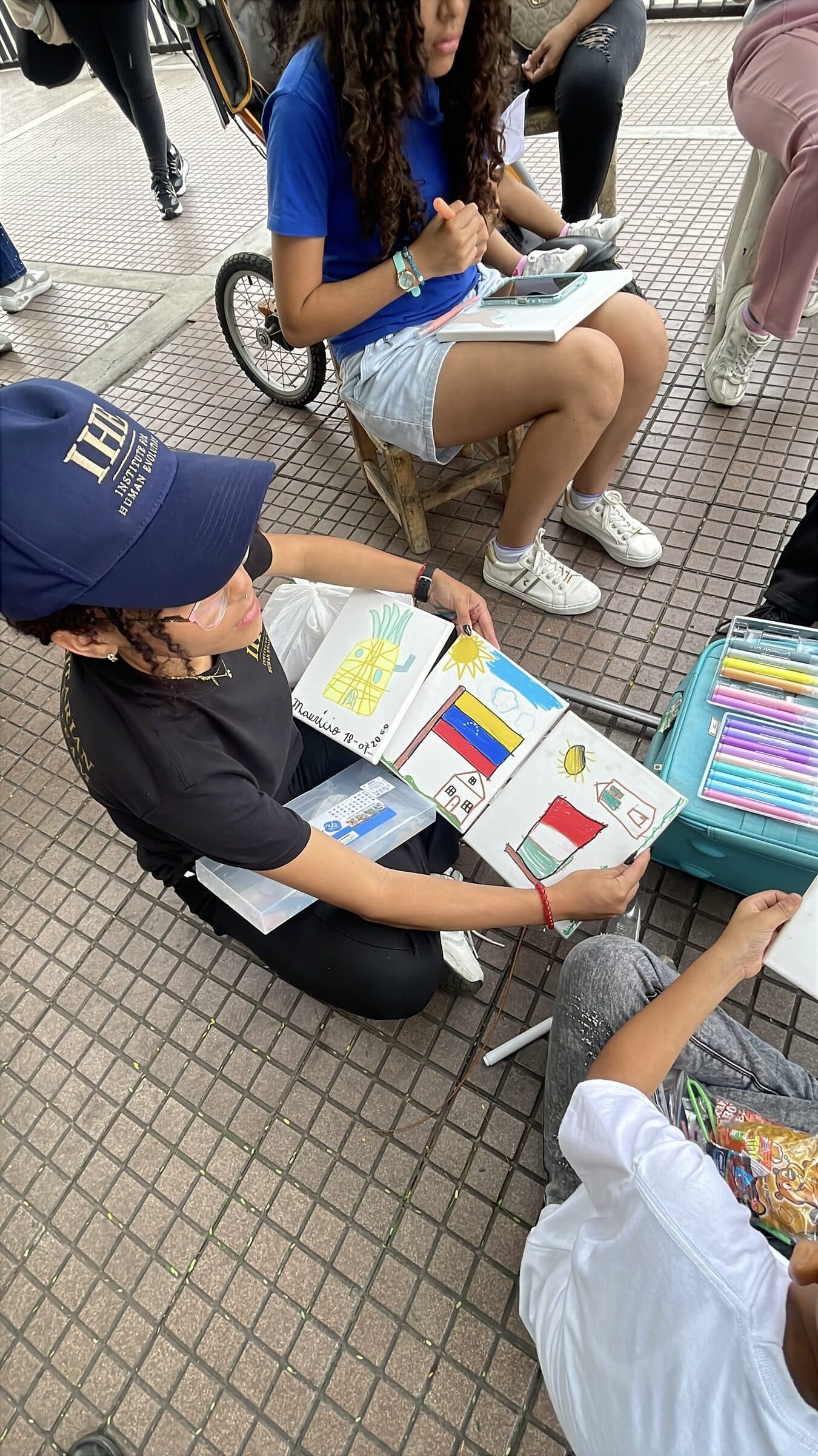 An IHE volunteer in a team cap holds up three finished canvases painted by children, including a Venezuelan flag and a pineapple.
