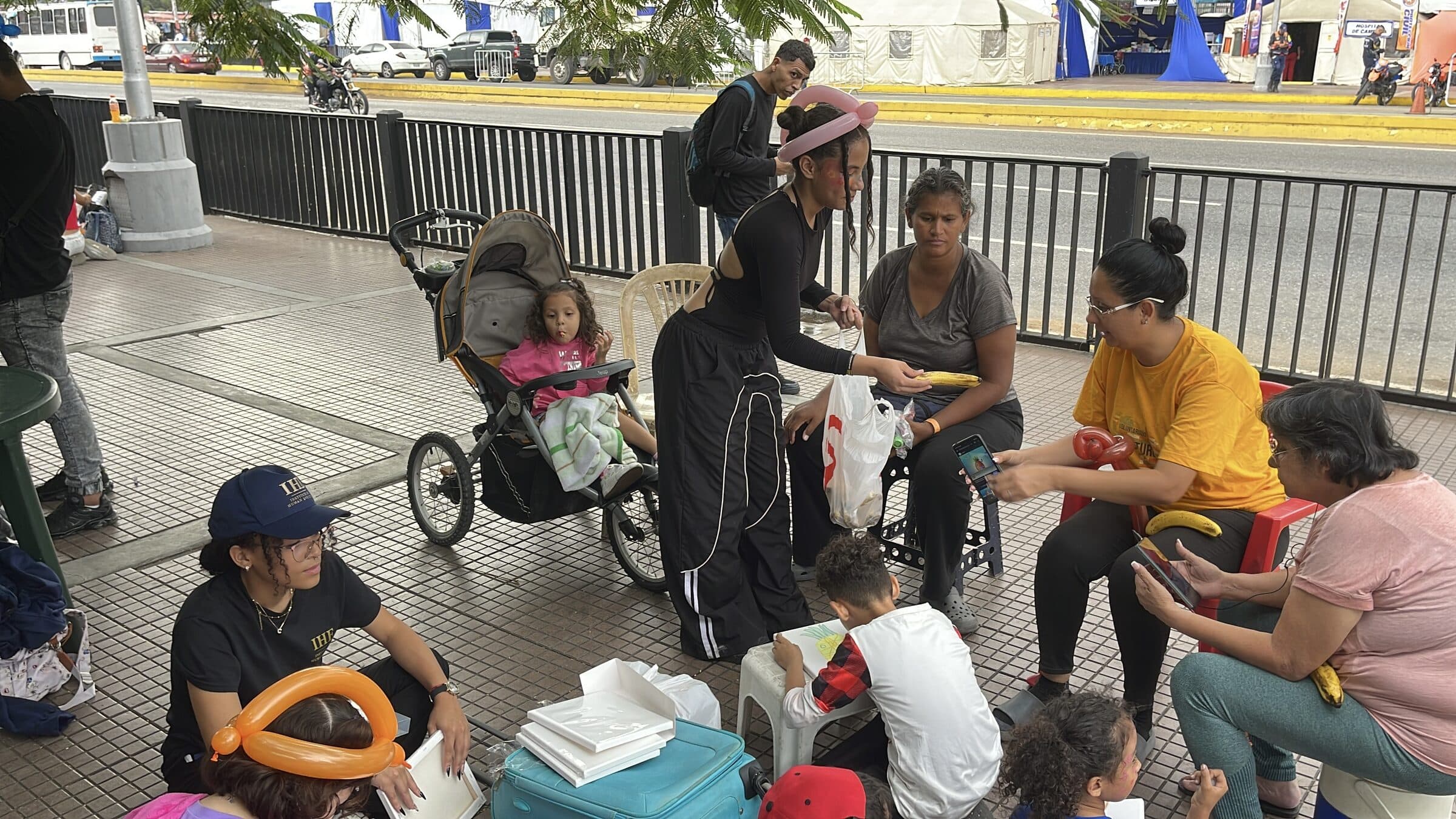 A volunteer hands fresh fruit to a mother beside her child's stroller; the camp's tents are visible across the street in Caracas.