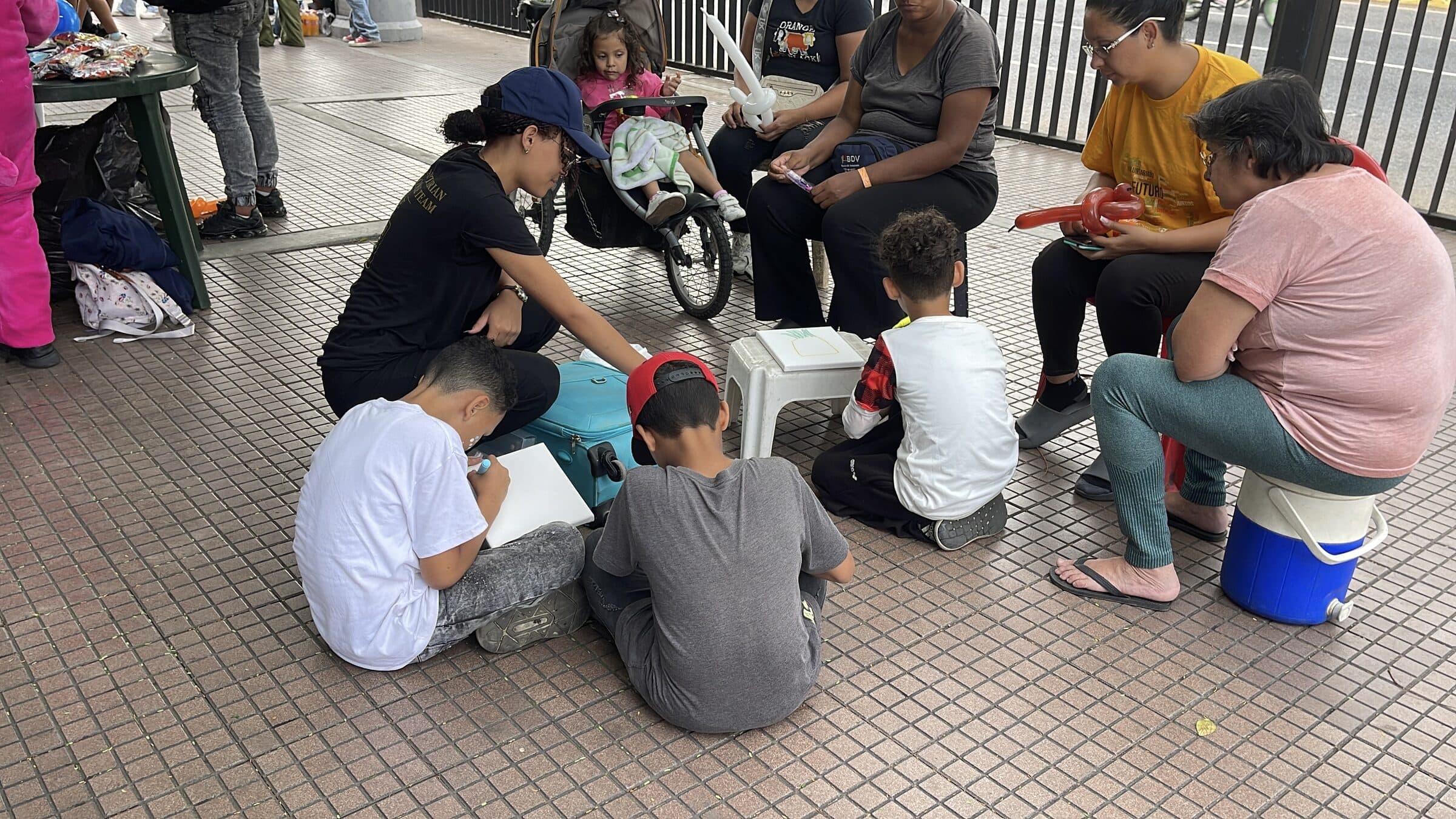 An IHE Humanitarian Response Team volunteer leads a drawing circle with children and their mothers on a sidewalk next to the camp.