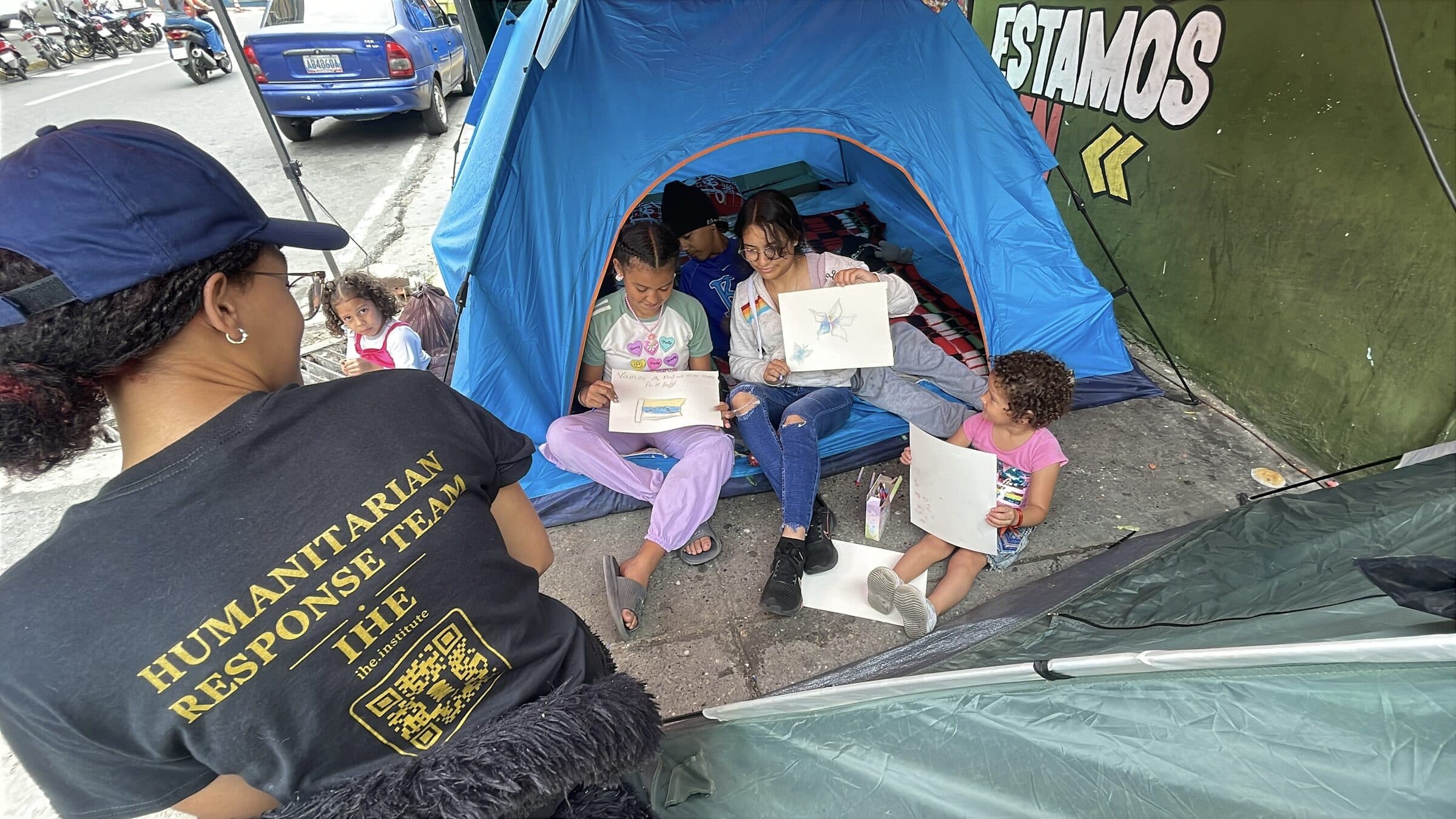 Children proudly show their finished drawings, including a Venezuelan flag, beside an IHE volunteer at the camp art workshop.