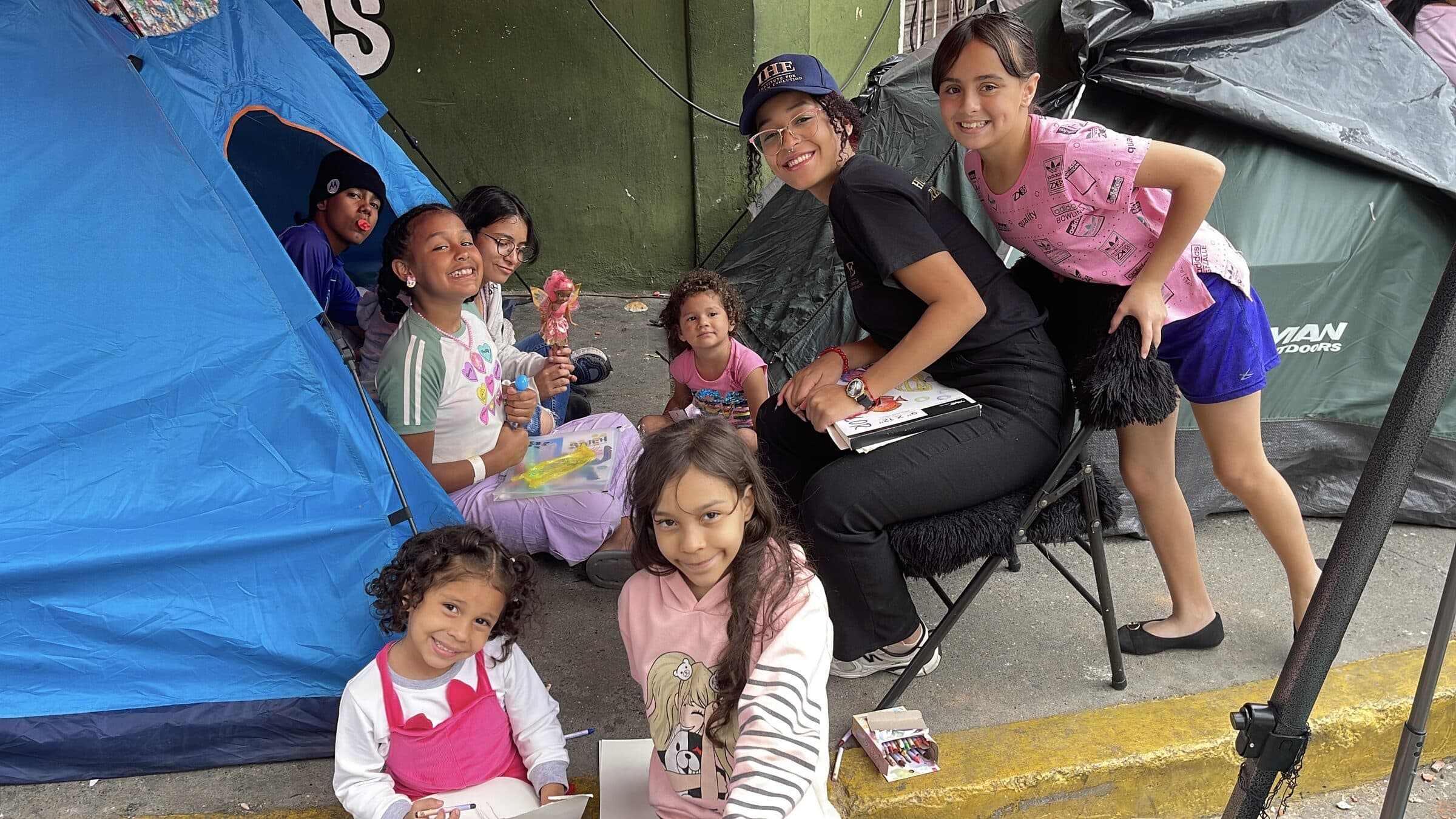 An IHE volunteer surrounded by smiling children in front of the tents at the camp, a spontaneous group portrait during the July mission